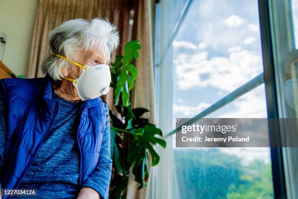 wide angle low shot distraught elderly senior caucasian woman looking out the window feeling loneliness wearing an n95 protective face mask to prevent the spread of covid sars ncov 19 coronavirus swine flu h7n9 influenza illness during cold and flu season - compromised immune system stock pictures, royalty-free photos & images