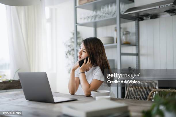 young asian woman drinking coffee and having breakfast, talking on smartphone while working from home in the living room using laptop computer - social responsibility stock pictures, royalty-free photos & images