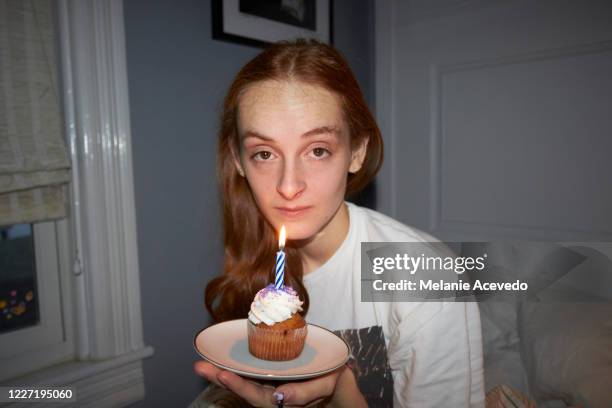 teenage girl in her bed holding a birthday cupcake with a candle on it. she is looking at the camera. she has long red hair. - cupcakes girls stock-fotos und bilder