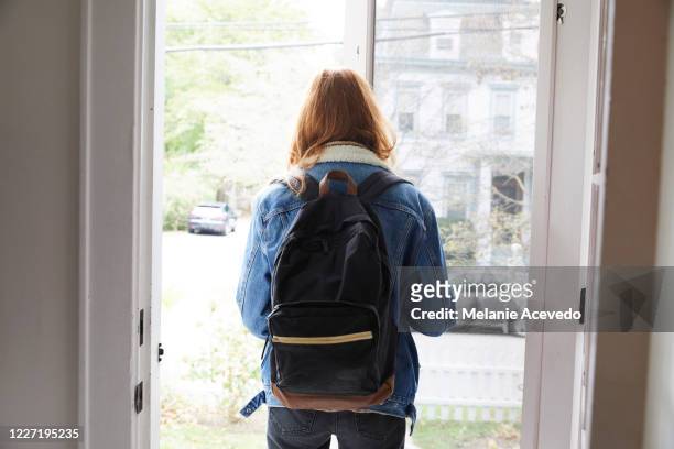 teenage girl walking out the front door of her house. back view of her leaving the house. she is on her way to school, wearing a back pack and holding the door open. - aussteigen stock-fotos und bilder
