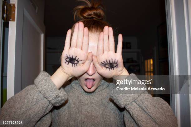 portrait of a teenage girl covering her eyes with her hands which have drawings of eyes on them. shot from the chest up. indoors. she is sticking out her tongue. - hand uitsteken stockfoto's en -beelden
