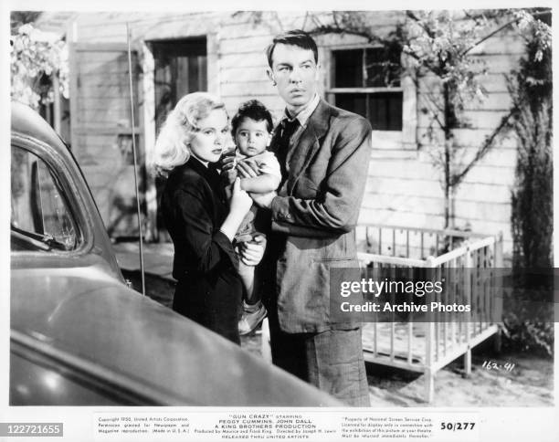 Peggy Cummins stands holding baby with male actor in a scene from the film 'Gun Crazy', 1950.