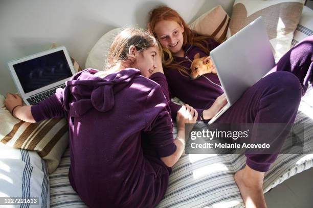 two teenage girls wearing matching purple sleep onesies and lying on a couch playing on their computers. - bodysuit stock pictures, royalty-free photos & images