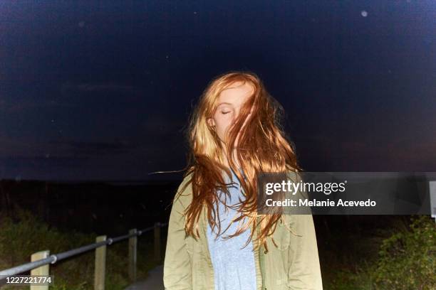 young girl on the beach at night with her long red hair blowing across her face. the sky is deep blue behind her. flash photo. - head in sand stock-fotos und bilder