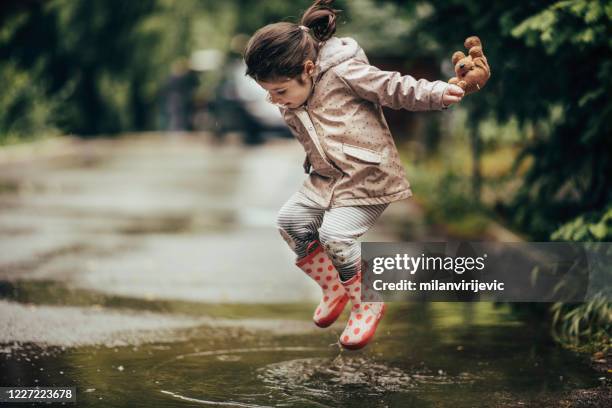 petite fille de sourire jouant dans une photo de stock de flaque d’eau - kway photos et images de collection