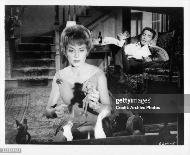 Janet Blair holding wooden sculpture while Peter Wyngarde is passed out on the couch in a scene from the film 'Burn, Witch, Burn', 1962.