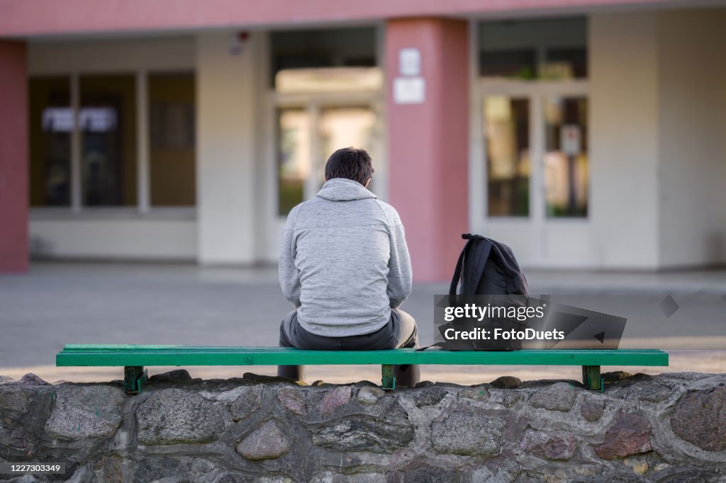 One young man sitting on bench at school yard. Break time. Back view.