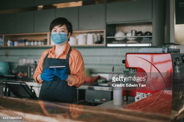 eine asiatische chinesische kleinunternehmerin mit gesichtsmaske und schutzhandschuhen übergibt das dessert an ihre kundin an der küchentheke lächelnd mit blick auf die kamera - essenzielle berufe und dienstleistungen stock-fotos und bilder