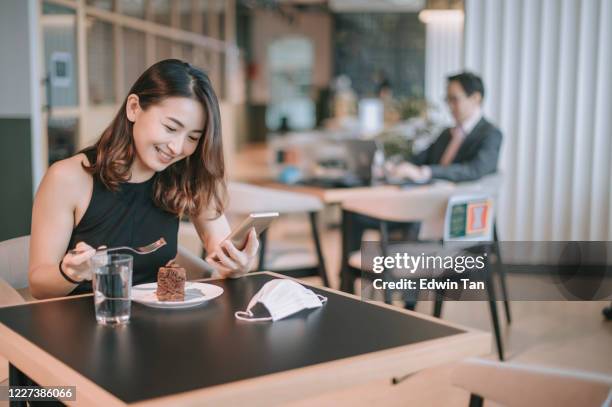 an asian chinese female enjoying her dessert chocolate cake during her tea break in cafeteria while practicing social distancing using her phone communicating with her friend - social distancing restaurant stock pictures, royalty-free photos & images