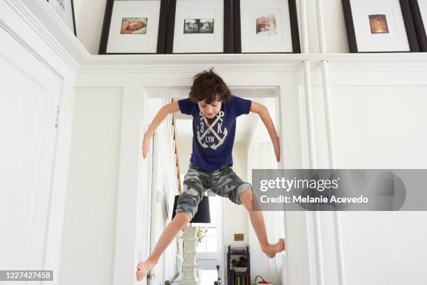 young boy with brown curly hair climbing up the door frame opening using hands and feet to climb the sides of the walls looking down at the ground beneath him low angle view picture frames mounted on the ceiling above his head - climbing frame stockfoto's en -beelden