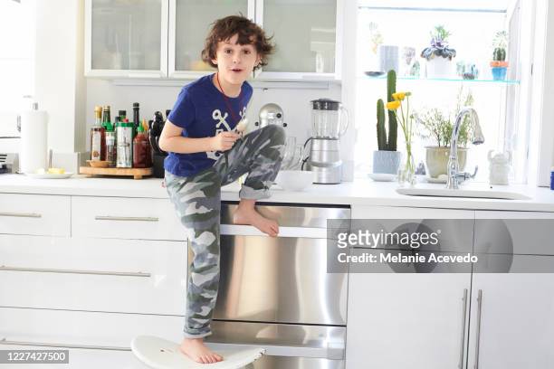 young boy with brown curly hair brown eyes sitting on the kitchen counter wearing dark blue graphic t-shirt and camouflage pants front leg dangling over the counter while the other leg is bent resting on the counter top - graphic t shirt stock-fotos und bilder
