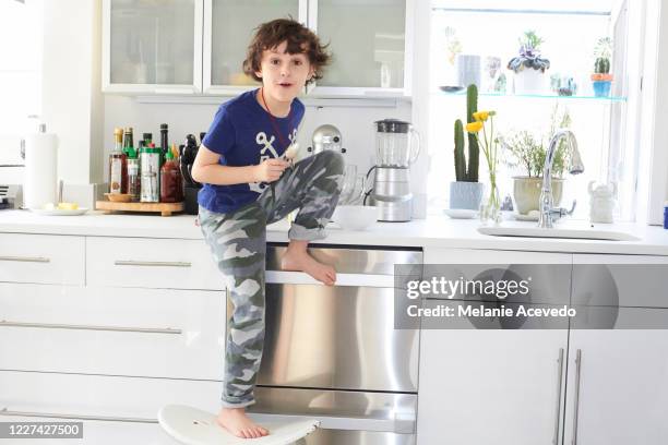 young boy with brown curly hair brown eyes sitting on the kitchen counter wearing dark blue graphic t-shirt and camouflage pants front leg dangling over the counter while the other leg is bent resting on the counter top - graphic t shirt stock pictures, royalty-free photos & images