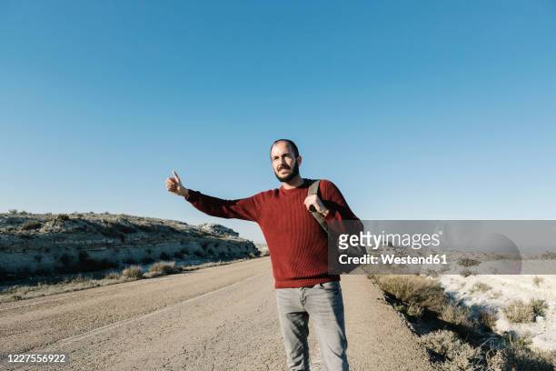 man gesturing while hitchhiking on roadside at desert against clear blue sky - hitchhike stock-fotos und bilder