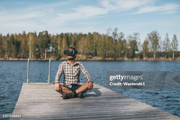 young man sitting on jetty, wearing vr glasses - eskapismus stock-fotos und bilder