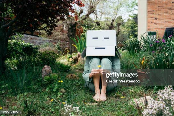 young woman with cardboard box on her head sitting barefoot in garden - schüchtern stock-fotos und bilder