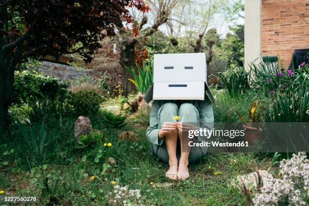 young woman with cardboard box on her head sitting barefoot in garden - embarrassment stock-fotos und bilder