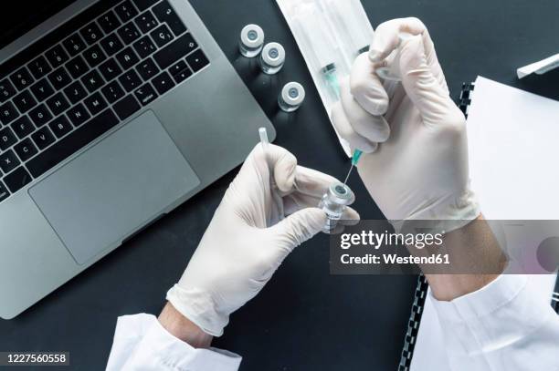 cropped image of doctor's hand injecting syringe in vial over laptop at laboratory desk - spiral notebook table stock pictures, royalty-free photos & images