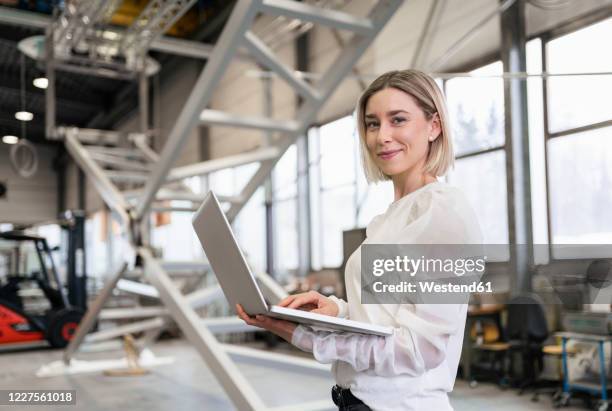 portrait of smiling young woman using laptop in a factory - portrait im raum stock-fotos und bilder