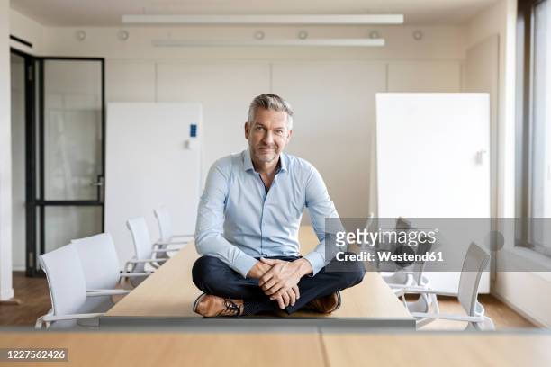 portrait of confident mature businessman sitting on table in conference room - cross legged stock pictures, royalty-free photos & images