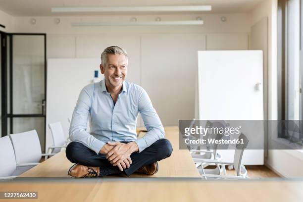 portrait of smiling mature businessman sitting on table in conference room - cross legged stock pictures, royalty-free photos & images