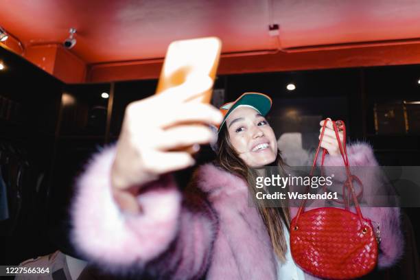 happy woman taking selfie through smart phone while wearing fur jacket and holding leather purse in store - handtasche stock-fotos und bilder