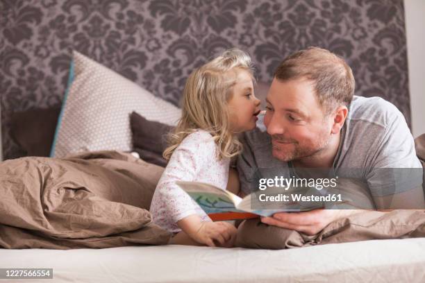 father and daughter reading a book in bed, whispering in ear - child whispering stock-fotos und bilder