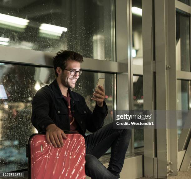 smiling young man with suitcase sitting at rainy window using smartphone - airport rain stock-fotos und bilder