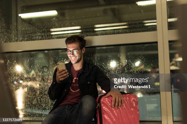 smiling young man with suitcase sitting at rainy window using smartphone - airport smartphone stock-fotos und bilder