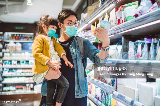 young asian dad & his lovely little daughter, both with medical face masks doing grocery shopping for organic snacks for kids in supermarket - use by label bildbanksfoton och bilder