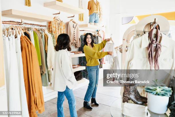 shop owner helping smiling woman choose outfit while shopping in clothing boutique - veleiding stockfoto's en -beelden