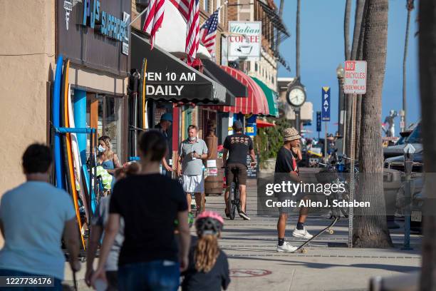 People walk and skateboard on the sidewalk past businesses on a summer day Monday, July 20, 2020 in Newport Beach, CA.