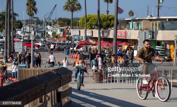 People walk, ride and skateboard on the sidewalk past businesses near the pier on a summer day Monday, July 20, 2020 in Newport Beach, CA.