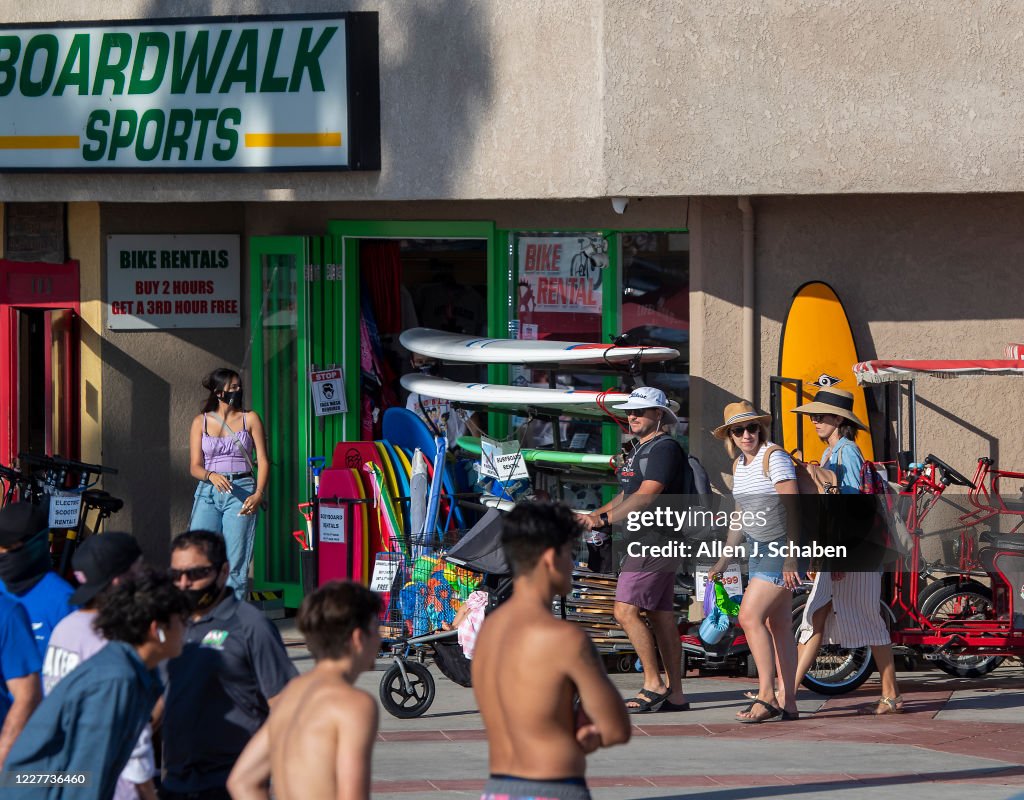 People walk and skateboard on the sidewalk past businesses on a summer day in Newport Beach