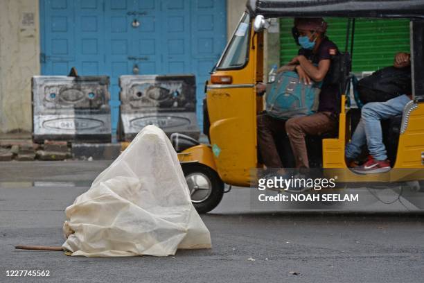 An auto-rickshaw passes by a homeless man sitting on a road under a metro rail bridge as he covers himself with a polythene sheet during a monsoon...