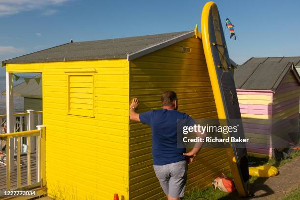 Man stands next to a bright yellow beach hut and waits patiently for his family to come along on the seafront promenade at Whitstable, on 18th July...