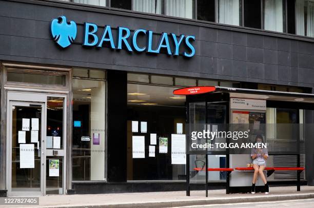 Commuter pedestrian wearing a face mask or covering due to the COVID-19 pandemic, sits inside a bus shelter by a branch of a Barclays bank in central...