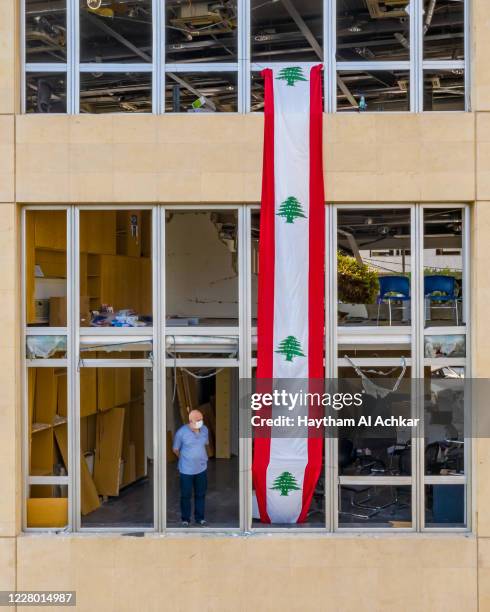 Man stands in the window of a damaged apartment building, draped with banner in the style of the Lebanese flag, on August 12, 2020 in Beirut,...