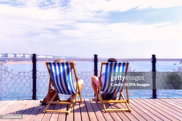 Couple seated on deckchairs enjoy the view of Brighton beach from the palace pier in East Sussex.