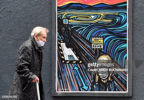 An elderly pedestrian wearing a face mask or covering due to the COVID-19 pandemic, walks past graffiti depicting the subjects within famous...