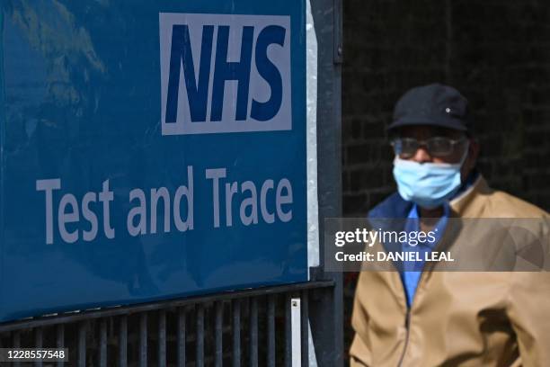 Man wearing a face mask or covering due to the COVID-19 pandemic, basses a sign for Britain's NHS Test and Trace service, as he leaves from a novel...