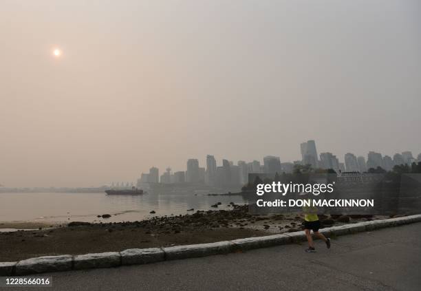 Person jogs along the Stanley Park seawall in Vancouver, Canada, with a view of the skyline under a heavy smog from clouds and smoke due to forest...