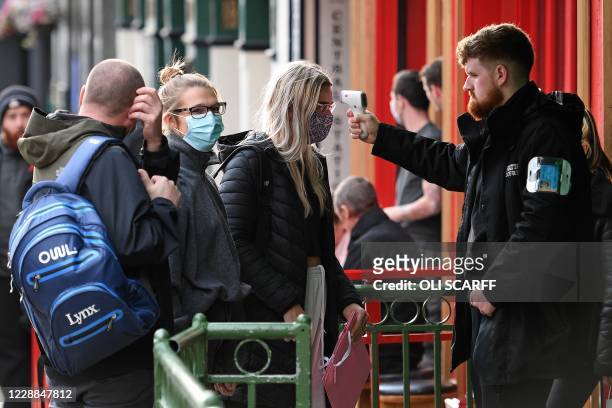 Security guard uses a handheld thermometer to take the temperature of customers, wearing face masks or coverings due to the COVID-19 pandemic, as...