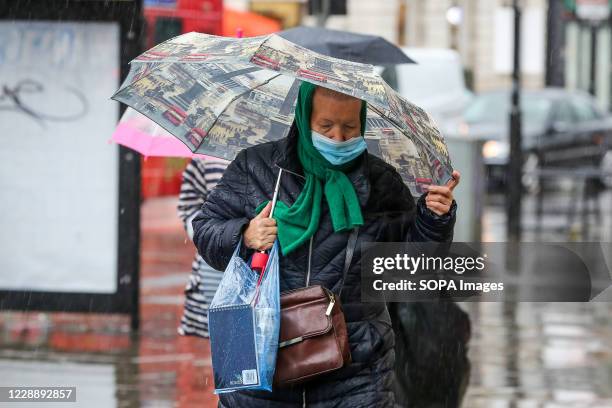 An elderly woman wearing a face mask shelters from the rain underneath an umbrella on a wet and windy day caused by Storm Alex.