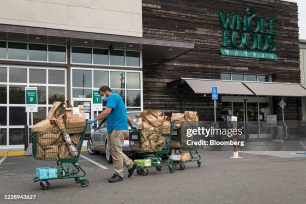 An independent contractor wearing a protective mask and gloves moves shopping carts with Amazon Prime grocery bags to a car outside a Whole Foods...