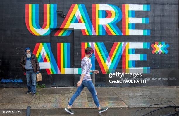 Pedestrian wearing a face mask or covering due to the COVID-19 pandemic, walks past a mural reading "U Are Alive..... So get your head out of your...