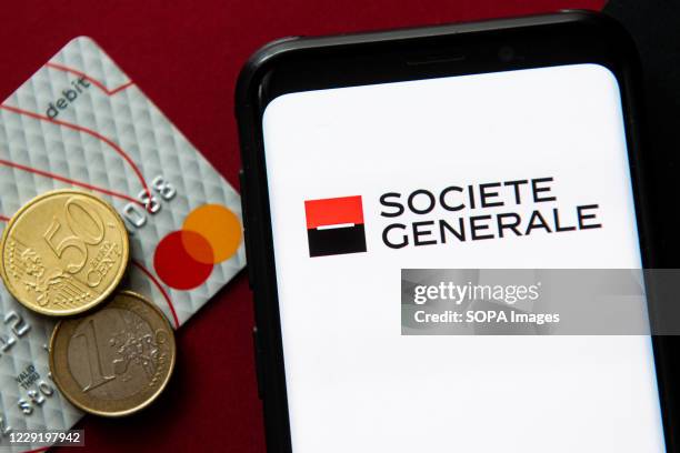 In this photo illustration a Societe Generale logo seen displayed on a smartphone next to a debit card and two euro coins.