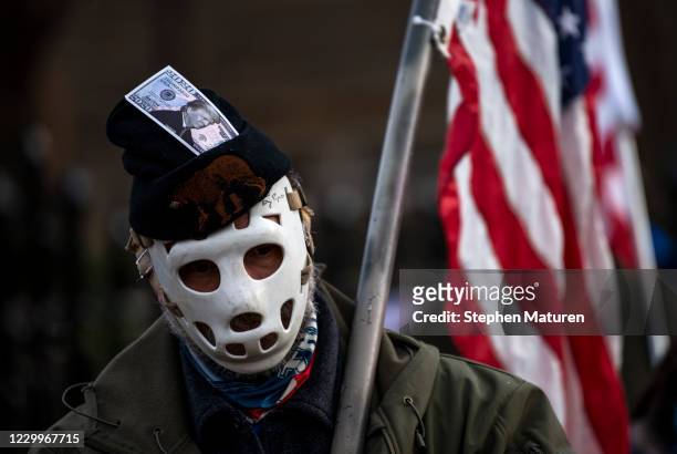 Man wears a hockey mask and a hat with a dollar bill imprinted with President Donald Trump looks on as protesters rally outside the Governor's...