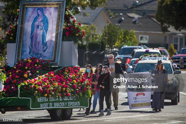 Handful of participants walking while saying the Rosary, followed by a long line of decorated vehicles participating in the annual Procession and...