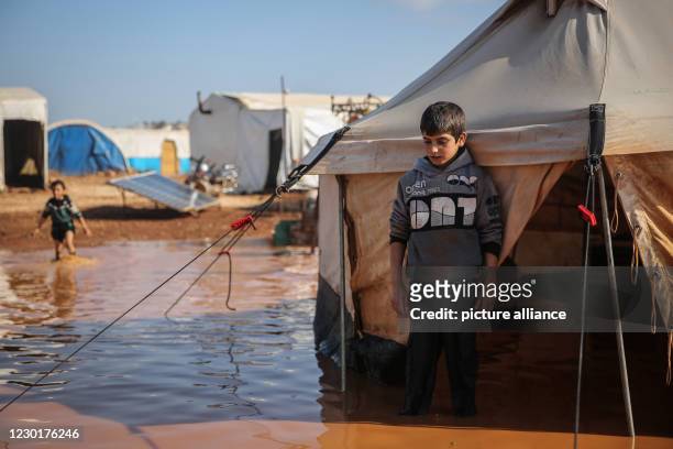 December 2020, Syria, Kafr Aruq: A Syrian boy stands amidst the muddy water outside his tent that has been flooded as a result of heavy rain at a...