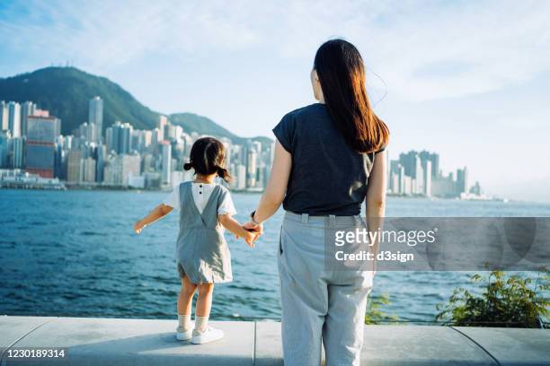 rear view of young asian mother holding hands with cute little daughter enjoying the sun by the promenade and looking over city skyline and victoria harbour on a lovely sunny day - business or women or family or travel stock-fotos und bilder